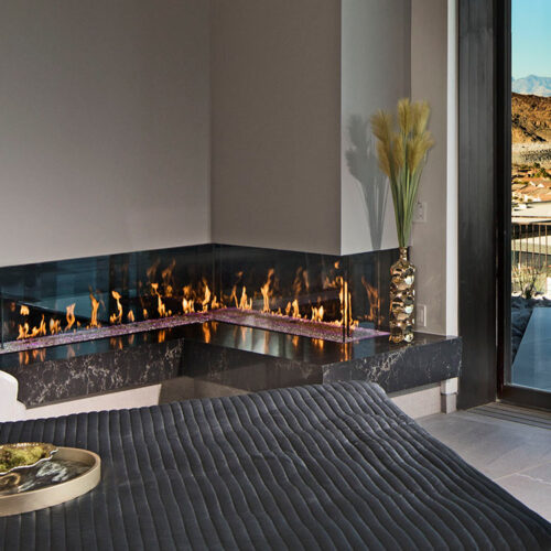 Modern bedroom with an L-shaped gas fireplace and large windows that provide a scenic mountain view.