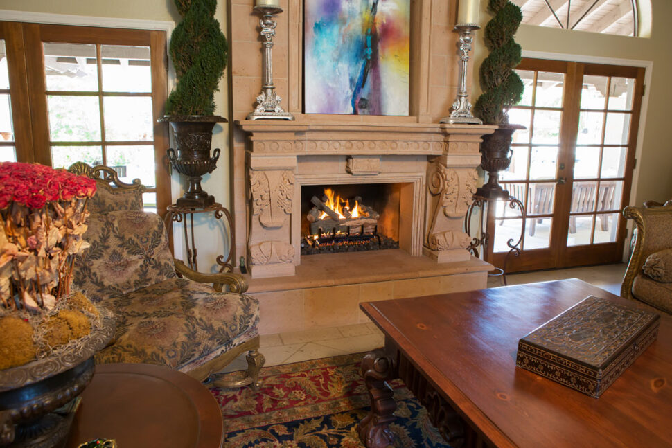 A cozy living room with an ornate fireplace, classic furniture, and natural light from large French doors.