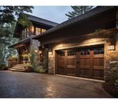 A stone and wood-clad home with two wooden double garage doors, illuminated by exterior lights, with a paved driveway and surrounded by lush greenery.