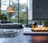 A modern living room featuring a sleek electric fireplace with vibrant flames, surrounded by contemporary decor, a large rug, and floor-to-ceiling windows that offer a view of lush greenery outside.