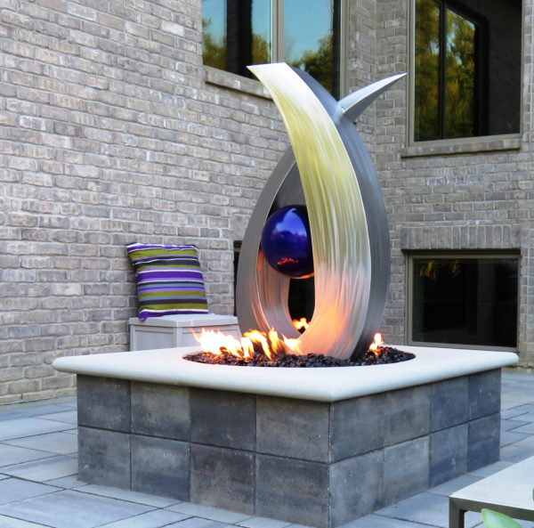 **Alt Text:** Modern outdoor fire feature with a sculptural design, featuring a silver and gold abstract structure with a blue orb, surrounded by flames on a tiled patio.